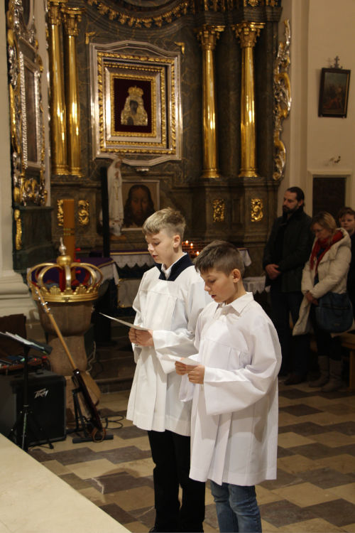 Dwóch młodych ministrantów w komżach, czytających z kartek. Stoją przed Ołtarzem.